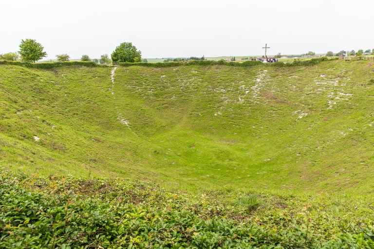 A crowd of people stand by a memorial cross, dwarfed by the sheer depth of the Lochnagarcrater.