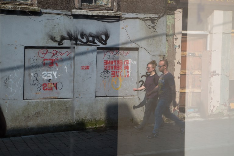 A man and a woman stride down Barrack Street, Cork City, caught in the reflection of an abandoned shop window