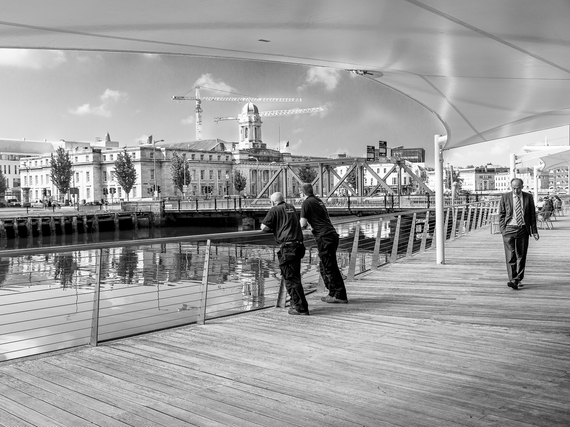 A suited man strolls along the boardwalk in Cork City while other workers take a break, leaning over the railing, cranes standing tall in the background.