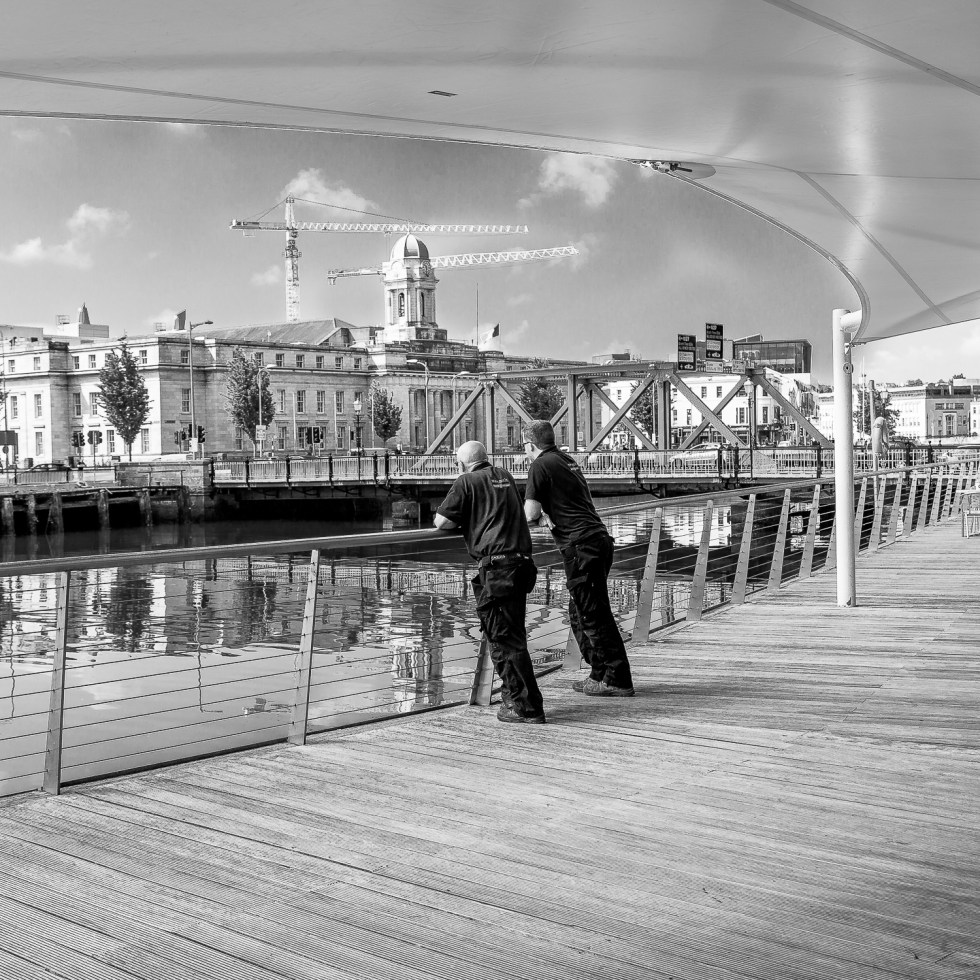A suited man strolls along the boardwalk in Cork City while other workers take a break, leaning over the railing, cranes standing tall in the background.