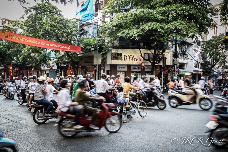 A cyclist walks her bicycle through a stream of mopeds in Hanoi, Vietnam